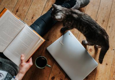 Foto van iemand die een boek leest op de grond met een laptop, koffie en een zwarte kat